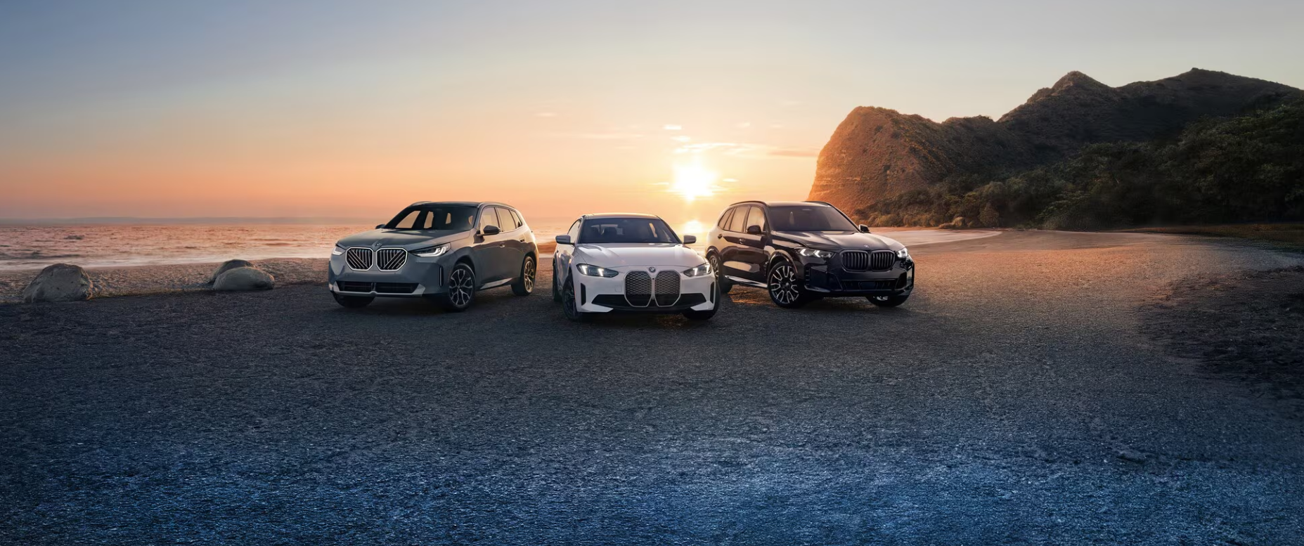 A trio of sleek BMW SUVs—silver, white, and black—parked on a pebbled beach at sunset with the ocean in background